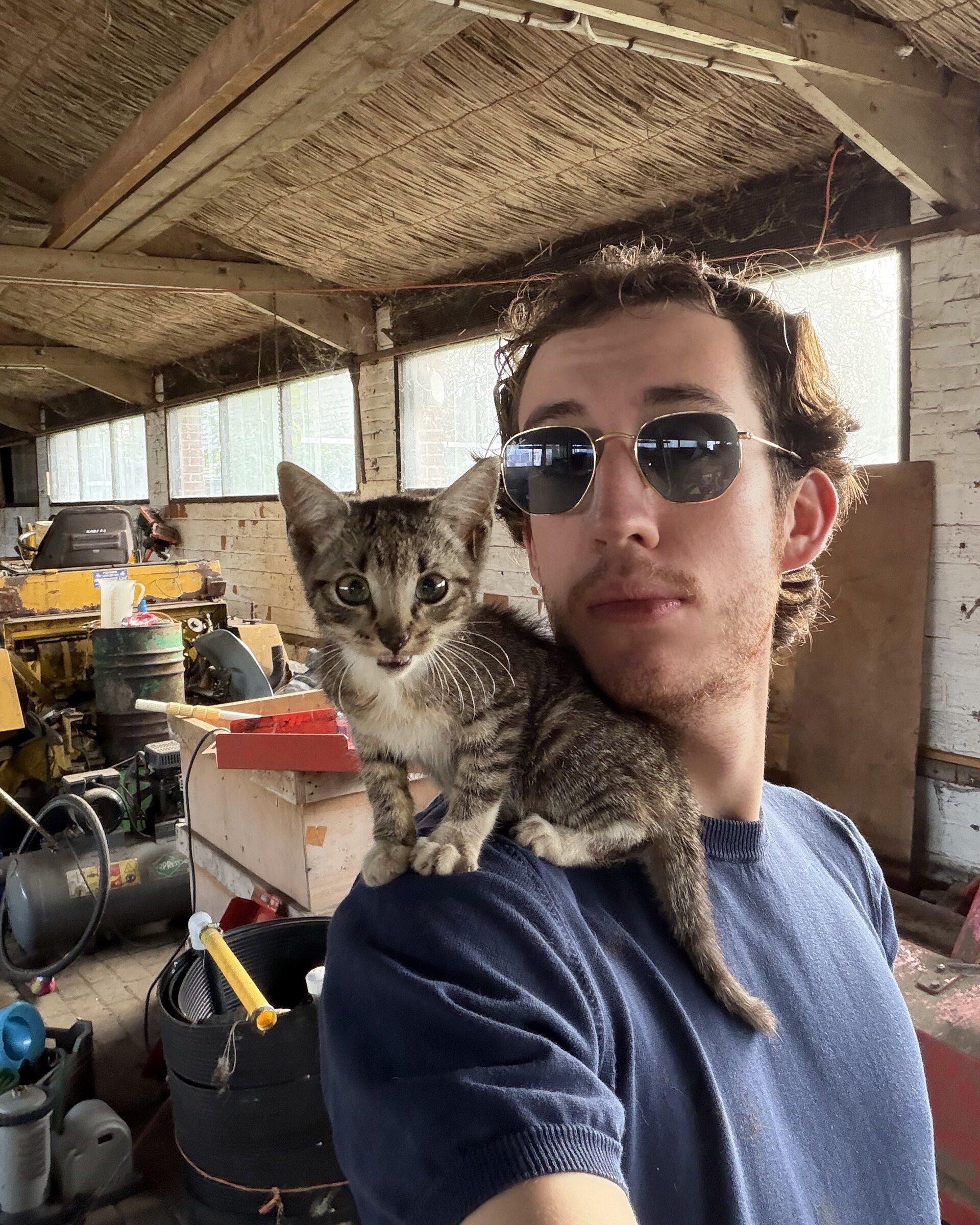 Selfie van Sven met zonnebril en een gestreepte kitten op zijn schouder in een schuur