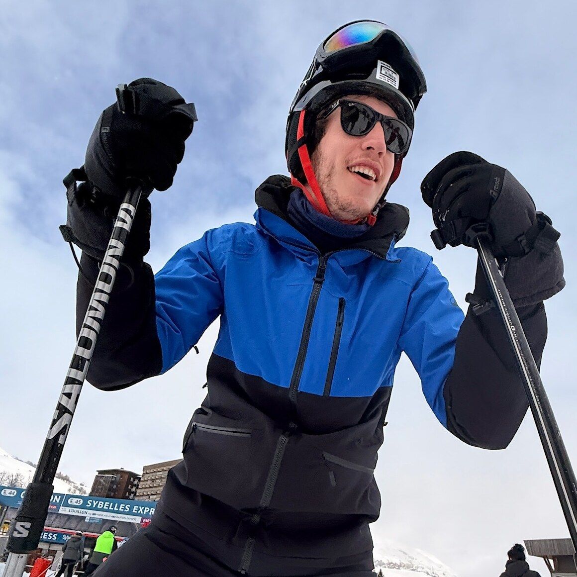 Sven lachend op de piste in skipak, met helm, zonnebril en skistokken in beide handen